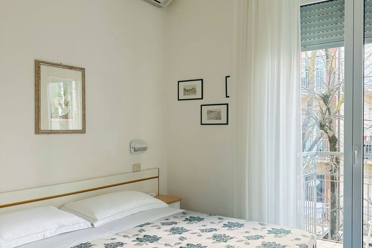 A bedroom with a bed, a ceiling fan, an air conditioning unit, a window with curtains, and a view of the outside.
