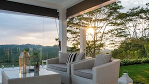 A modern outdoor seating area with wicker furniture and a glass table, overlooking a lush garden and distant hills. - Camelia Ranch (Baddegama)