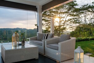 A modern outdoor seating area with wicker furniture and a glass table, overlooking a lush garden and distant hills.