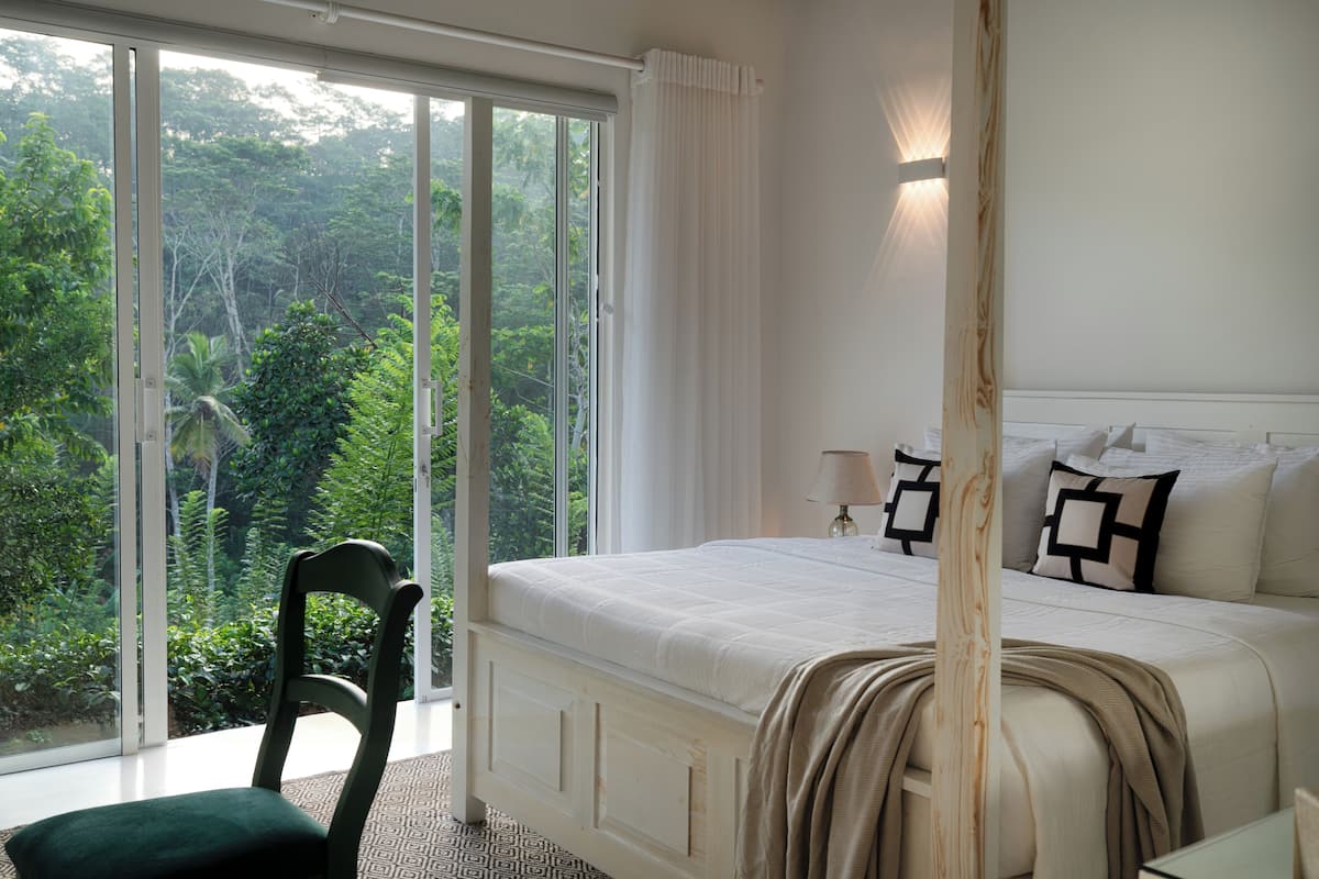 A bedroom with a large window, a bed with white bedding, and a view of greenery outside.