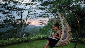 A person sitting in a wicker swing chair in a garden with a view of a sunset over a mountainous landscape. - Camelia Ranch (Baddegama)