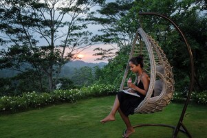 A person sitting in a wicker swing chair in a garden with a view of a sunset over a mountainous landscape.