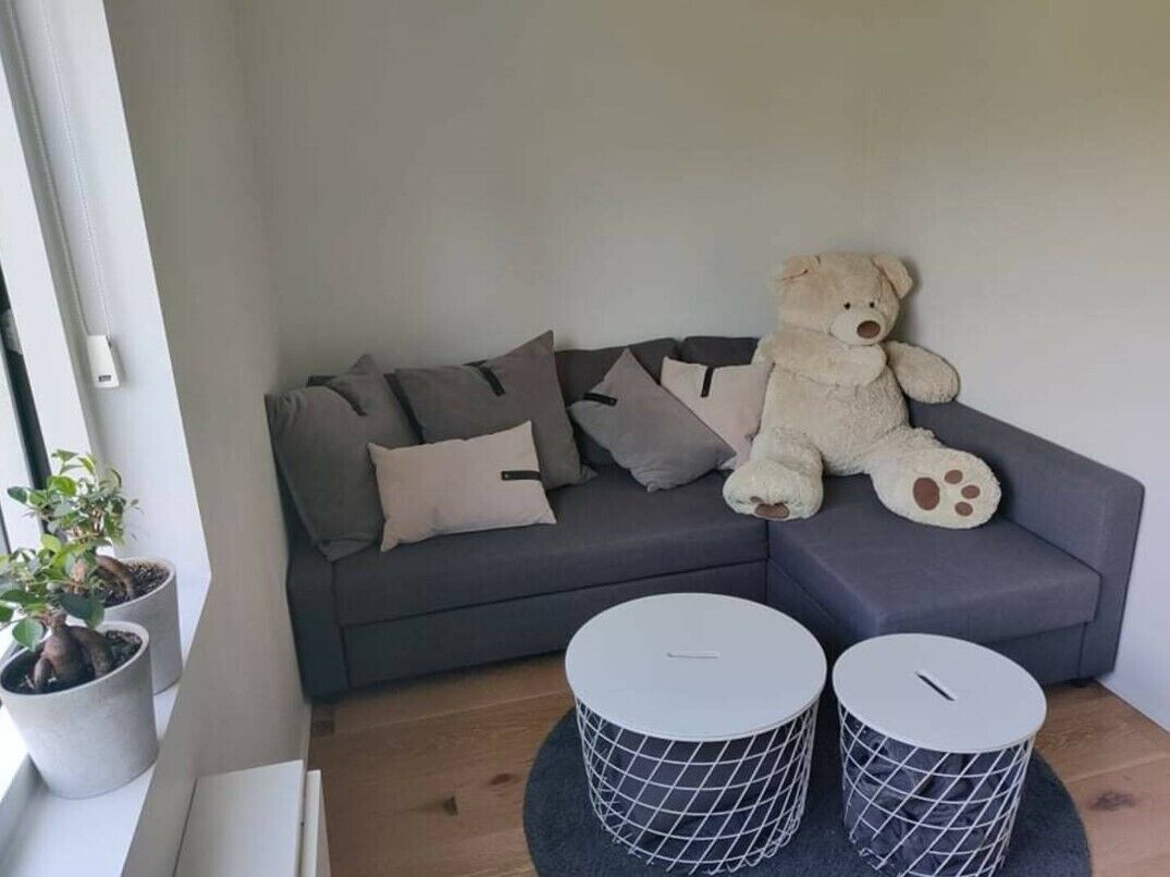 A grey sofa with multiple cushions, a large teddy bear, and two round wire baskets on a wooden floor.