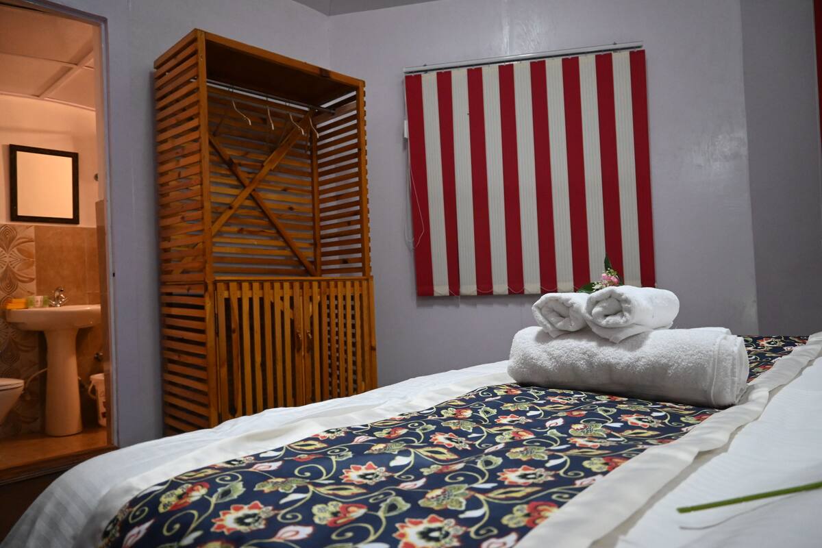 A hotel room with a bed, a wooden wardrobe, a striped wall hanging, and a bathroom with a sink and toilet.