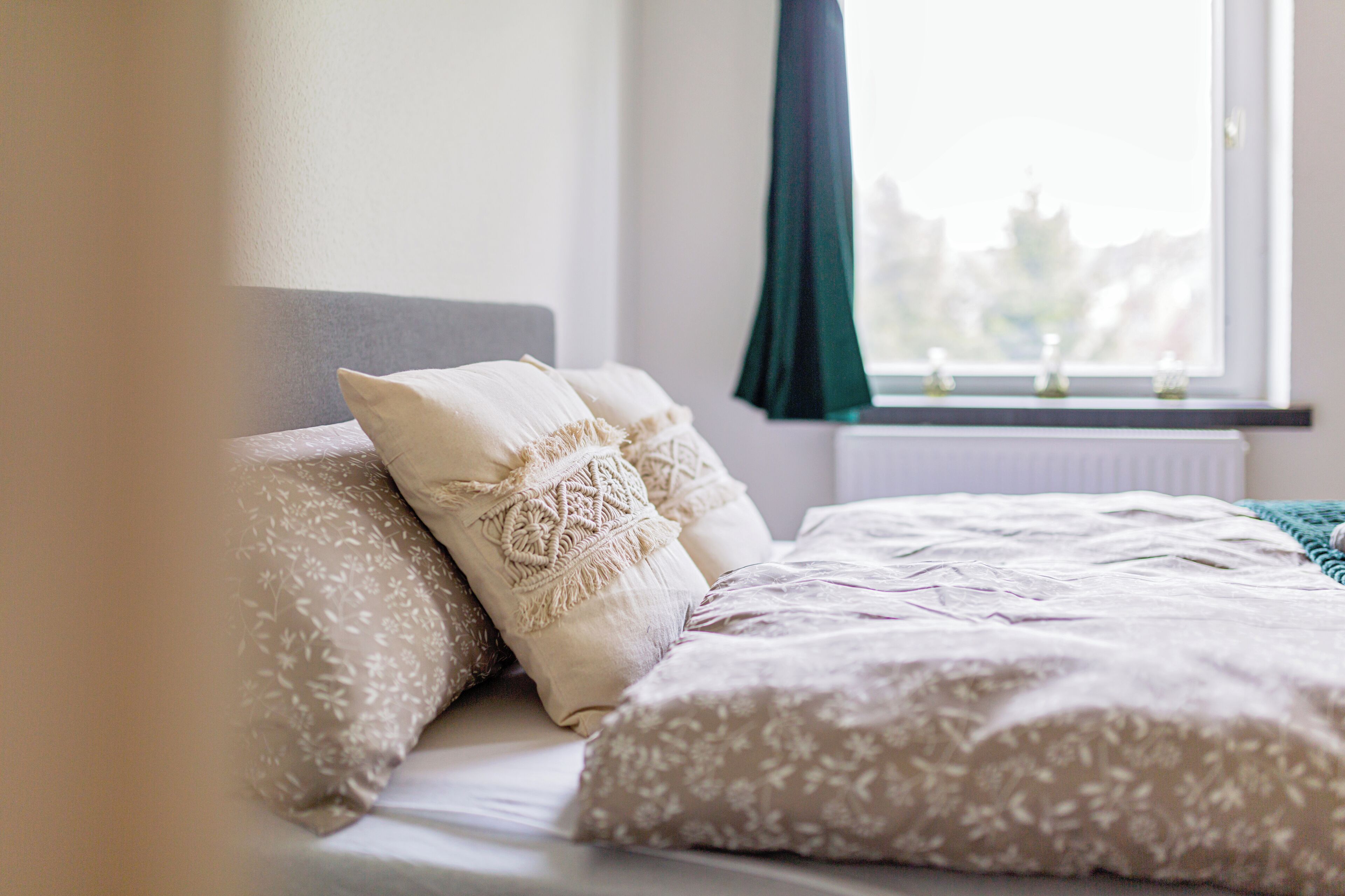 A neatly made bed with patterned bedding and pillows, a window with a view of greenery, and a radiator below.