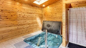 Interior de uma cabana de madeira com uma banheira de hidromassagem de vidro, um claraboia e uma cortina estampada.