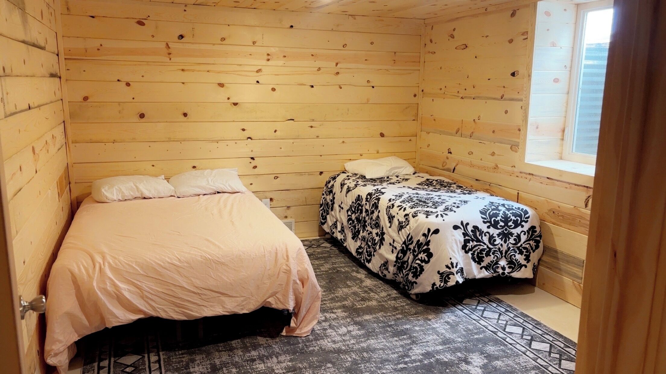 A small, cozy room with two beds, wooden walls, a window, and a patterned rug.