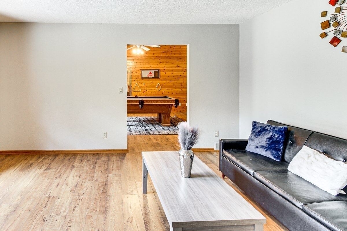 Ein Wohnzimmer mit Holzboden, einer schwarzen Ledersofa, einem Holztisch und Blick in einen Essbereich mit einer Holzwand und einem Billardtisch.