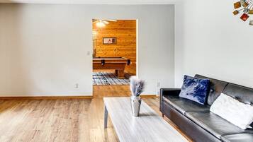 Ein Wohnzimmer mit Holzboden, einer schwarzen Ledersofa, einem Holztisch und Blick in einen Essbereich mit einer Holzwand und einem Billardtisch.