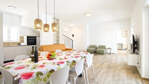 Egy modern nappali, étkezőasztallal, székekkel és egy kanapéval. A háttérben egy konyhasarok található, mikrohullámú sütővel és televízióval.