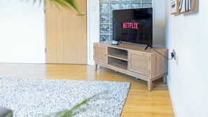 Een houten tv-meubel met een flatscreen televisie waarop het Netflix-logo staat, een getextureerd vloerkleed en een houten deur.
