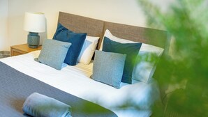 Een net opgemaakt bed met een grijs-witte deken, een blauw-witte kussenset, en een opgerolde grijze handdoek op een gertextureerd grijs laken.