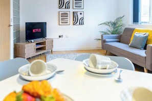 Ein modernes Wohnzimmer mit einem grauen Sofa, einem Couchtisch mit Tellern und Tassen, einem Fernseher, der 'NETFLIX' zeigt, und abstrakter Wandkunst.
