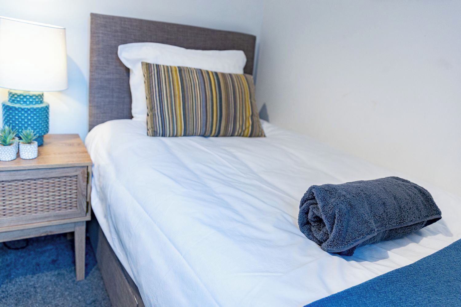 A neatly made bed with a striped pillow, a folded towel, and a small nightstand with a lamp and two potted plants.