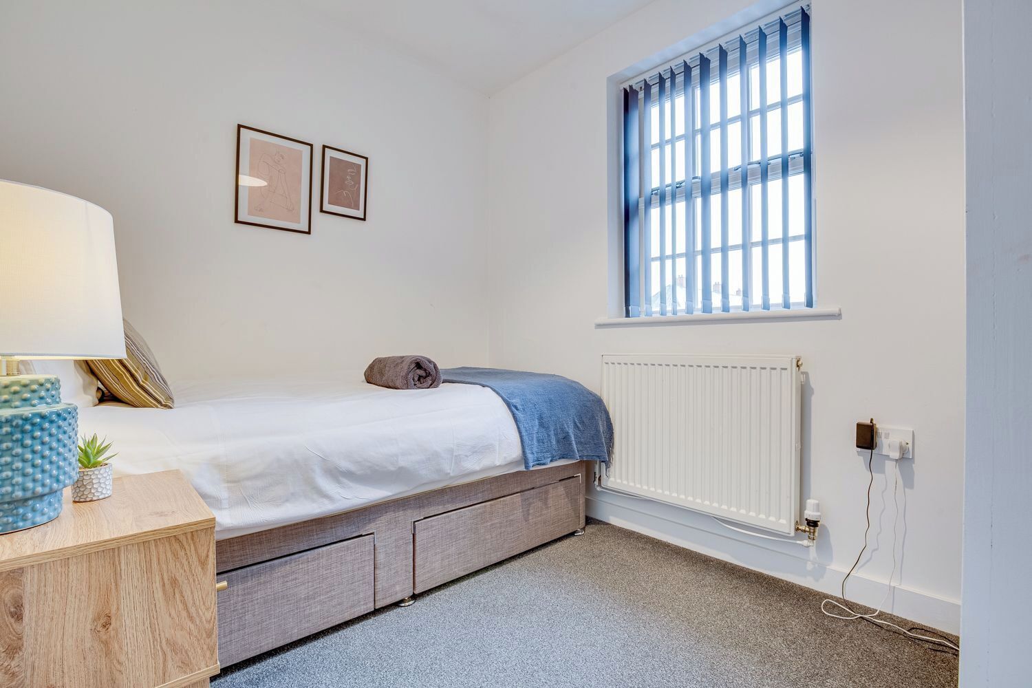 Une chambre à coucher avec un lit, une table de chevet avec une lampe, un radiateur et une fenêtre avec des stores.