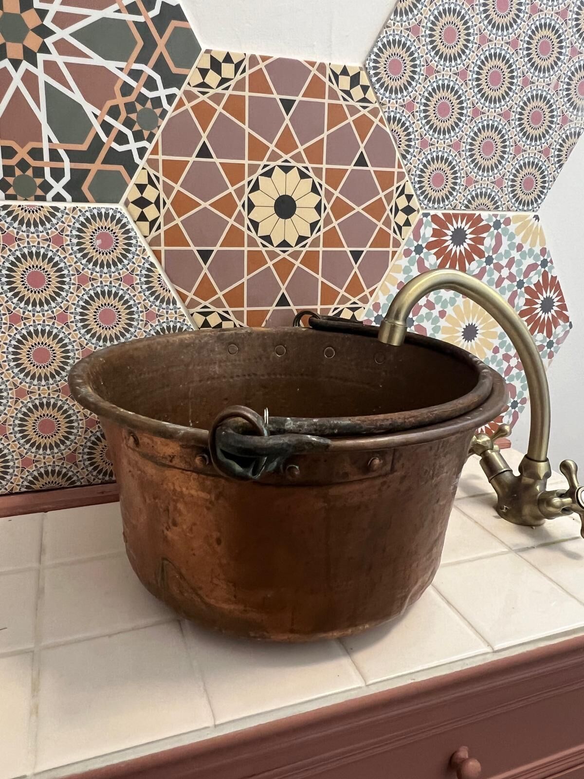 Een koperen wasbak met kraan, voor een muur met decoratieve tegels.