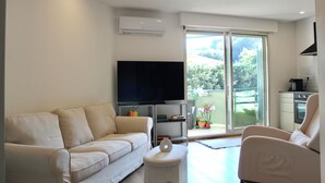 A modern living room with a sofa, a flat-screen TV, a small table, and a sliding glass door leading to a balcony with plants. - Charming apartment near the sea (Cavalaire-sur-Mer)