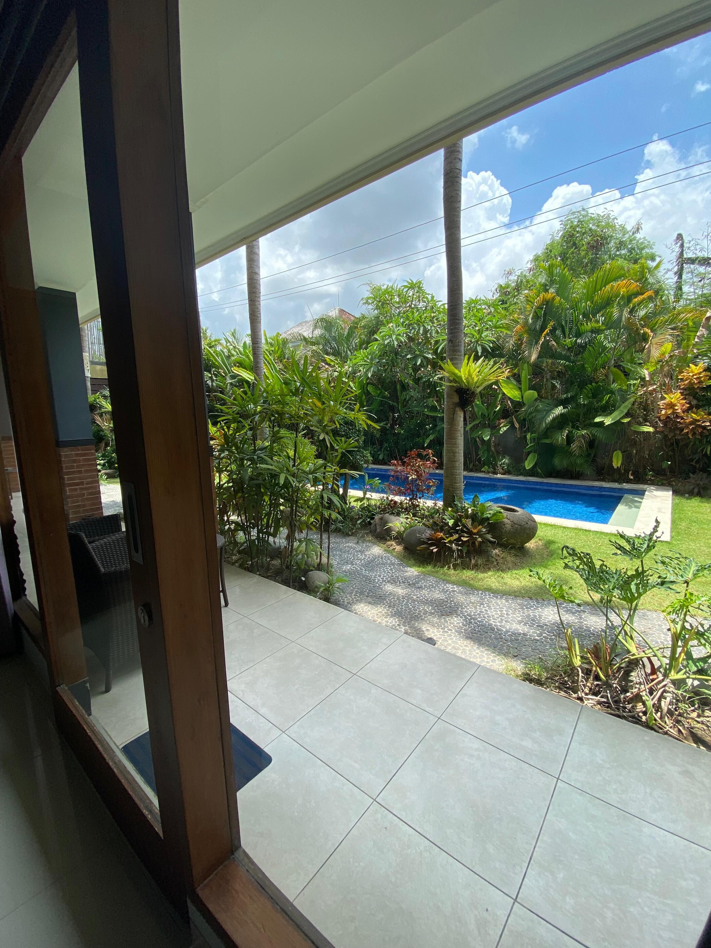 A view of a swimming pool surrounded by lush greenery, with a tiled patio area in the foreground.