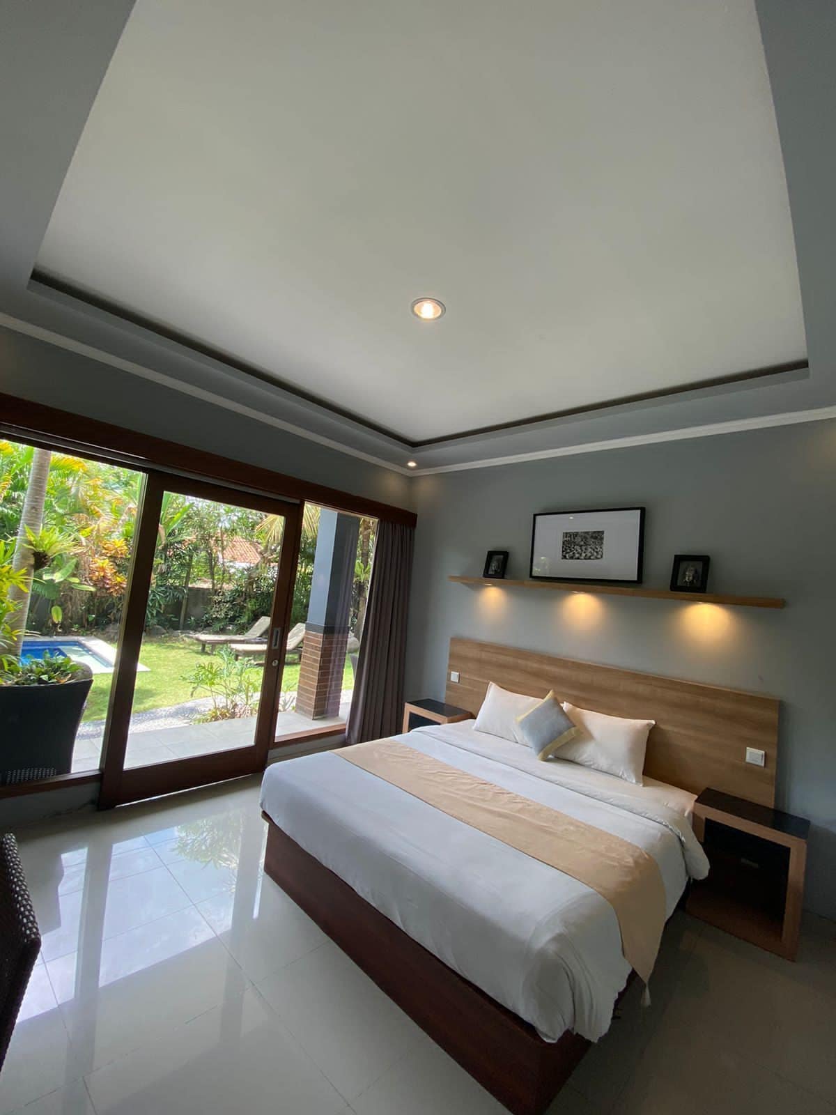 Une chambre moderne avec un grand lit, une tête de lit en bois, une table de chevet et une vue sur un jardin, accessible par une porte coulissante.
