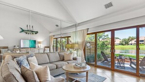 A spacious living room with a large sofa, a round coffee table, and a view of a garden through large sliding glass doors. - PlayaBella-Sandestin Escape|Lakefront|Golf Cart (Sandestin)