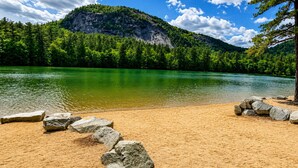 A lake with a sandy beach and rocks, surrounded by dense green forest and a mountain in the background. - Cranmore Retreat, Walk to Slopes, Hot Tub, Game Room, Dog-Friendly, Sleeps 16+ (Conway)