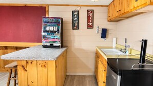 A compact kitchen with wooden cabinets, a countertop with a vending machine, a sink, and a refrigerator. - Cranmore Retreat, Walk to Slopes, Hot Tub, Game Room, Dog-Friendly, Sleeps 16+ (Conway)