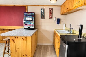 A compact kitchen with wooden cabinets, a countertop with a vending machine, a sink, and a refrigerator. - Cranmore Retreat, Walk to Slopes, Hot Tub, Game Room, Dog-Friendly, Sleeps 16+ (Conway)