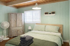 A bedroom with a bed, a wooden wardrobe, a standing fan, and a window with blinds.