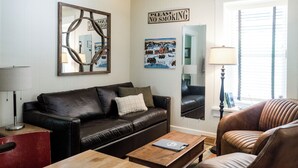 A living room with a leather sofa, a wooden coffee table, a brown armchair, a lamp, and wall art including a 'Please No Smoking' sign.