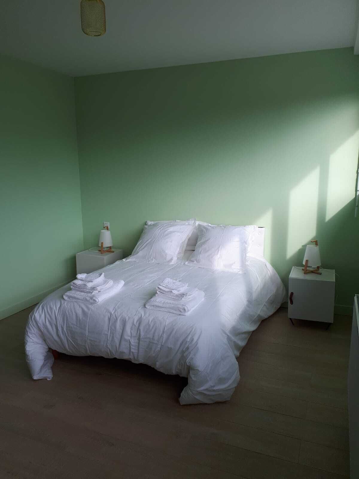 A neatly made bed with white linens, a small bedside table with a lamp, and a wall-mounted light fixture.