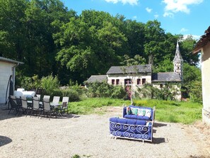 Eine Schotterterrasse mit Sitzgelegenheiten, ein rustikales Gebäude mit einem Turm und üppiges Grün im Hintergrund.