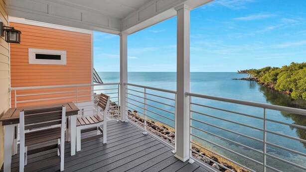 A covered outdoor area with a wooden table and chairs, overlooking a body of water and a rocky shoreline.