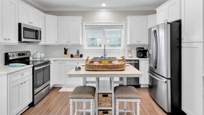 A modern kitchen with white cabinetry, stainless steel appliances, a central island with stools, and a window with blinds.