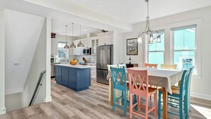 A modern kitchen with white cabinets, a blue island, stainless steel appliances, and a dining area with colorful chairs.