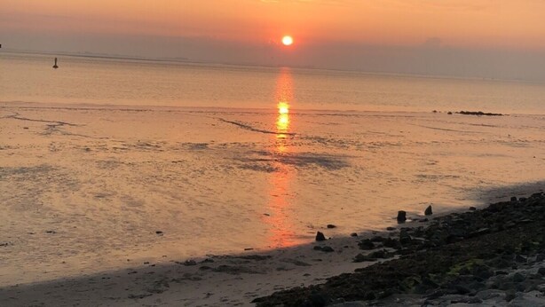 Auringonlasku kivikkoisella rannalla, jossa tyyni vesi heijastaa taivasta.