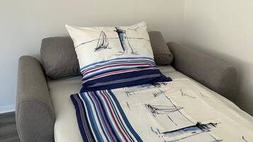 A single bed with a striped blanket and pillows, a framed picture above it, and a grey couch to the left.