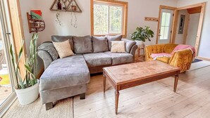 En stue med en grå hjørnesofa, en gul lænestol, et træsofabord og et stort vindue, der lukker naturligt lys ind.
