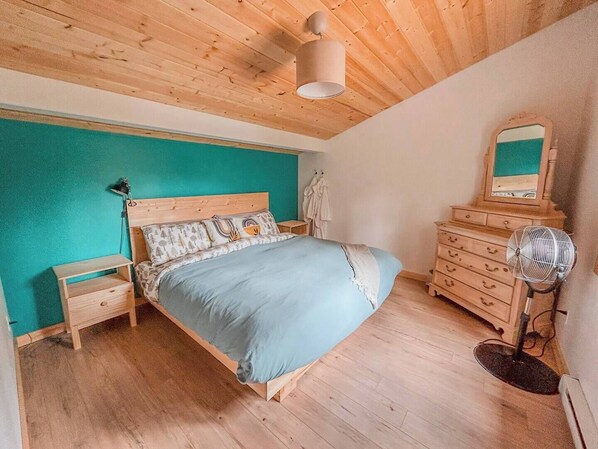 Een slaapkamer met een houten plafond, een bed met opvallende kussens, een nachtkastje, een dressoir en een ventilator.