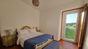 A bedroom with a bed, bedside tables, a bench, a hanging light fixture, and a view of a green landscape through a door. - Herdade do Vidigal (Montemor-o-Novo)