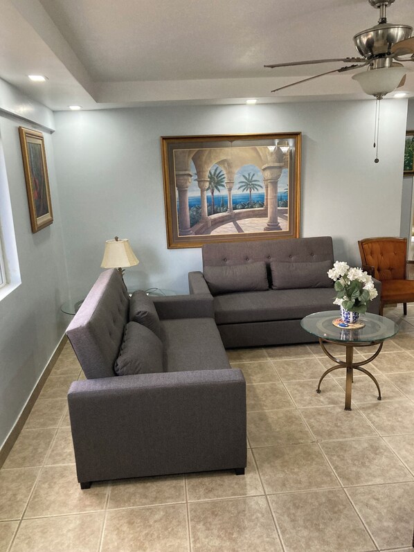 A living room with a sectional sofa, a glass coffee table, a painting of a tropical scene, and a ceiling fan. - Turtle Beach Towers - in Ocho Rios. (Ocho Rios)