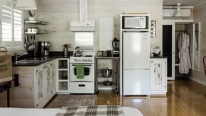 Cocina compacta con muebles blancos, estufa, microondas y refrigerador. Hay un estante pequeño con vajilla y una tetera sobre la estufa.