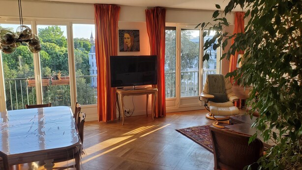 Ein großzügiger Wohnbereich mit großem Fernseher, bequemen Sitzgelegenheiten und einem Balkon mit Blick auf Grünflächen und Gebäude.