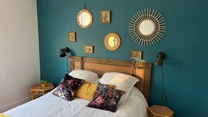 Ein Schlafzimmer mit einer türkisblauen Wand, einem Holz-Kopfteil, einem Bett mit weißer Bettwäsche und bunten Kissen, einem kleinen Holzschemel und Wandlampen.