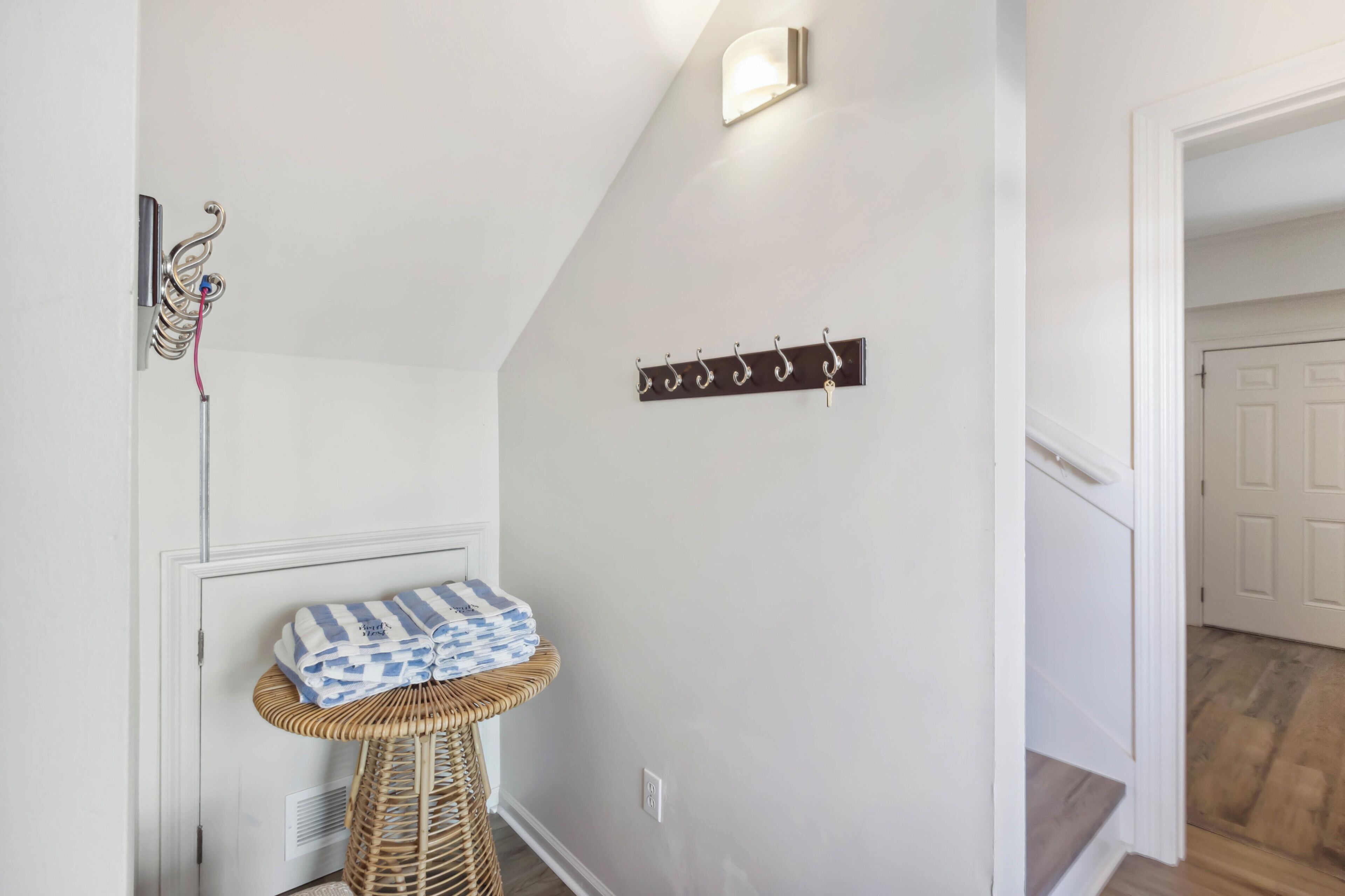 A hallway with a coat rack, a wall-mounted light, and a small wicker table with a towel.