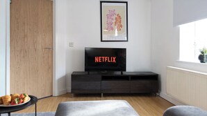 A modern living room with a dark wood floor, a grey sofa, a small round table with pastries, a dark wooden TV stand with a Netflix screen, and a framed botanical print on the wall. - Quaint 1 Bedroom 1st Floor Flat (Doncaster)