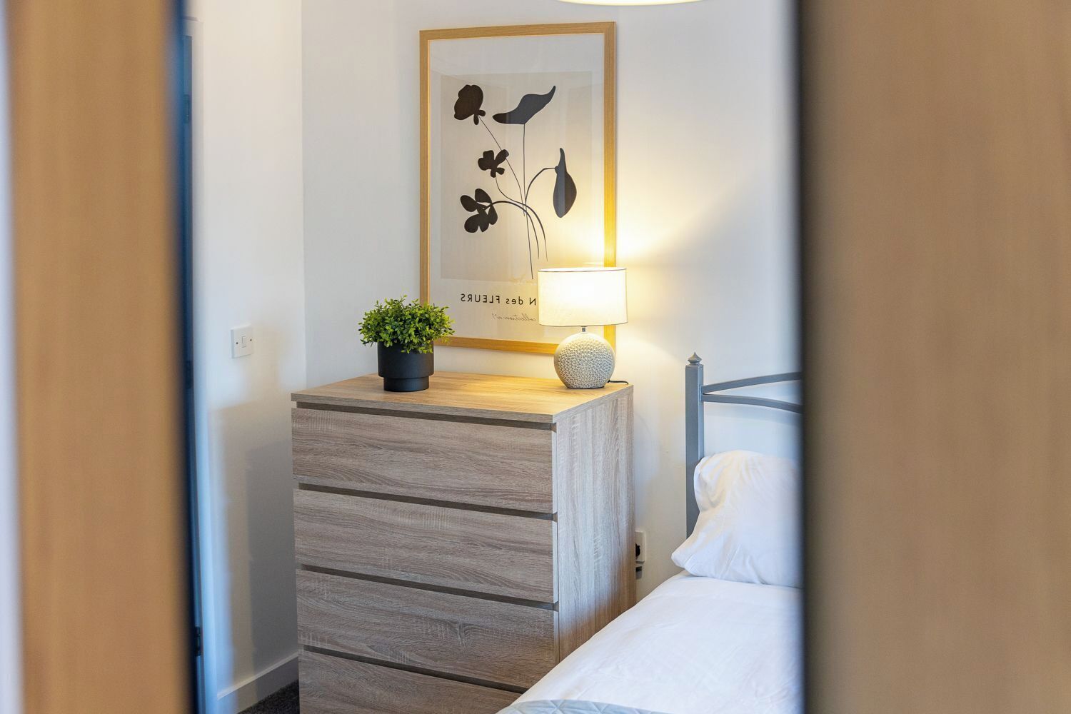Un dormitorio ordenado con cama, una mesita de noche con lámpara, una obra de arte enmarco y una planta en maceta.