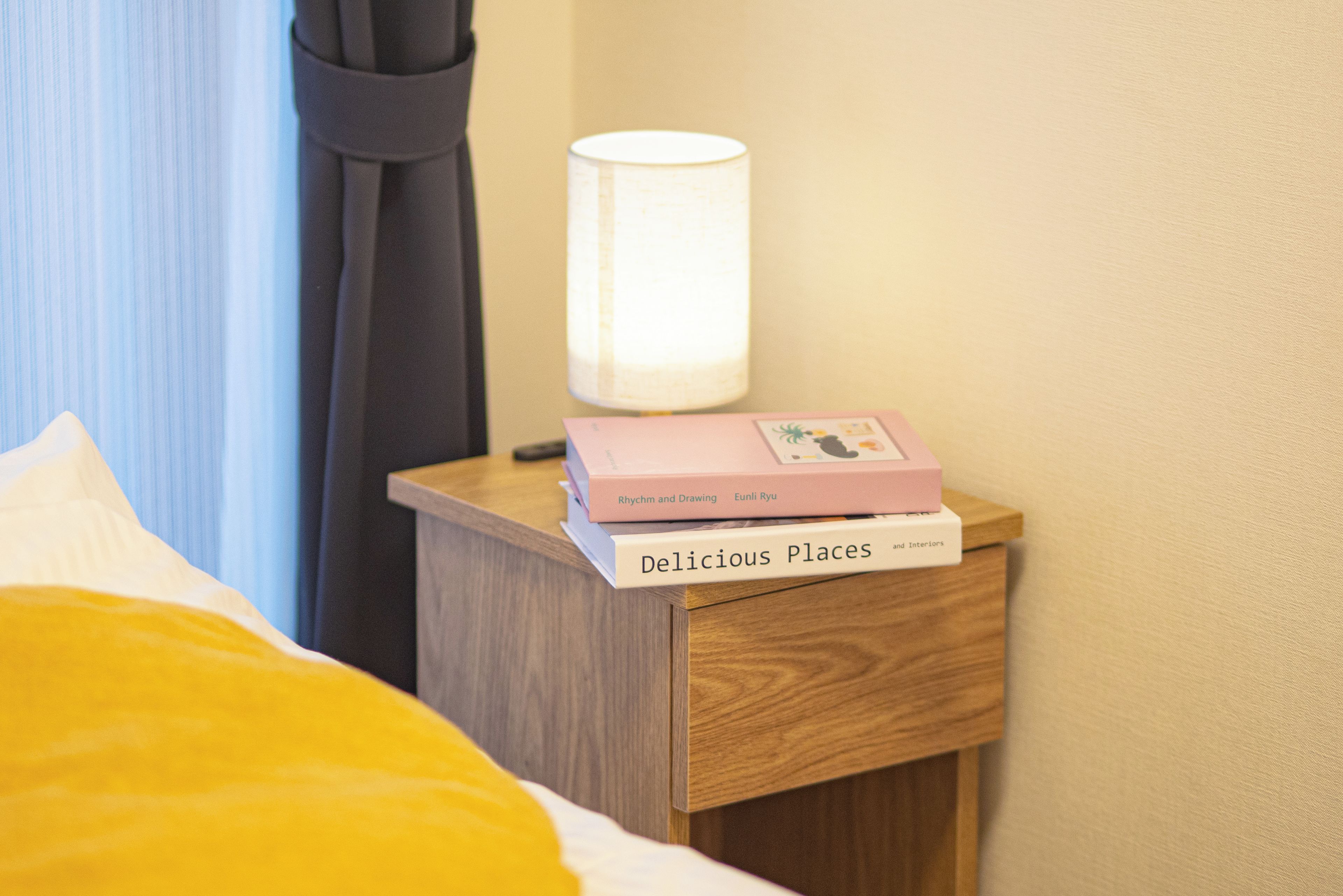 Een nachtkastje met een lamp en twee boeken, een bed met een gele deken, en een raam met gordijnen.