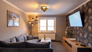 Una sala de estar con un sofá gris, una mesa de centro de madera, una lámpara de pie, un televisor en la pared y una ventana con persianas.