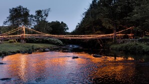 Un pont suspendu illuminé, enjambant une rivière et entouré d’arbres.