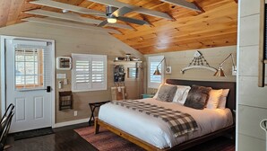 Une chambre avec un plafond en bois, un lit recouvert d’une couverture à carreaux, une table de chevet, un bureau et une fenêtre avec des stores.